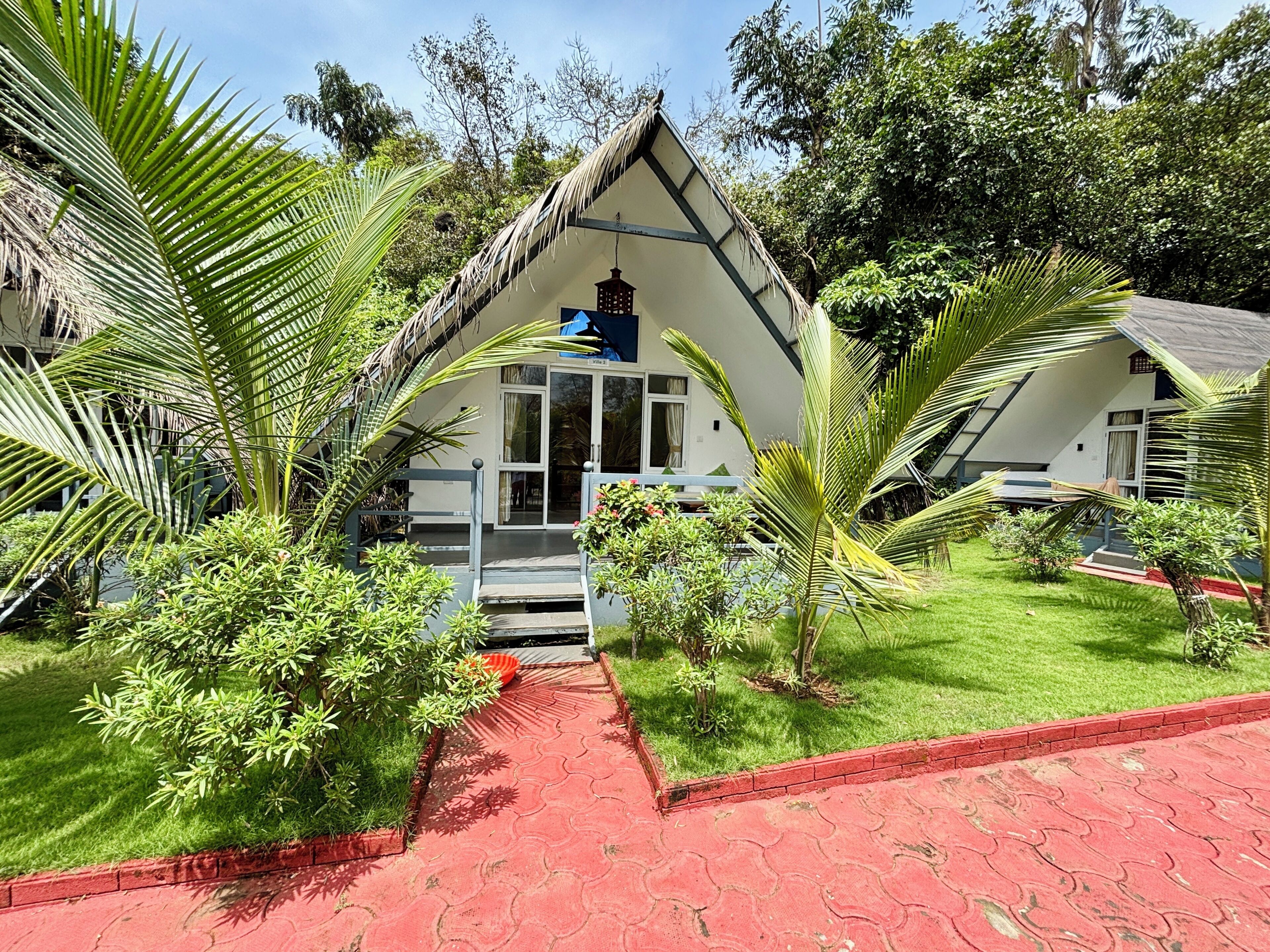 Un bungalow doté d’un toit de chaume, entouré d’une végétation luxuriante et dont l’entrée est accessible par un chemin rouge.