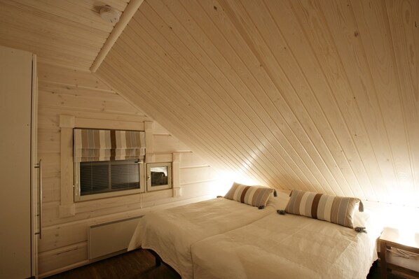A bedroom with a sloped ceiling, a bed with striped bedding, a window with blinds, and a radiator. - Ounasvaara Lakituvat chalets (Rovaniemi)