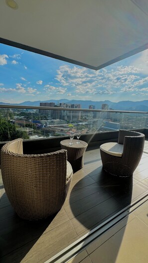A balcony with wicker furniture and a glass railing overlooking a cityscape with mountains in the distance. - El Poblado! 2hab+Studio+Balcony+ parking, AC, fast WIFI, The Best in Medellin! (Medellín)