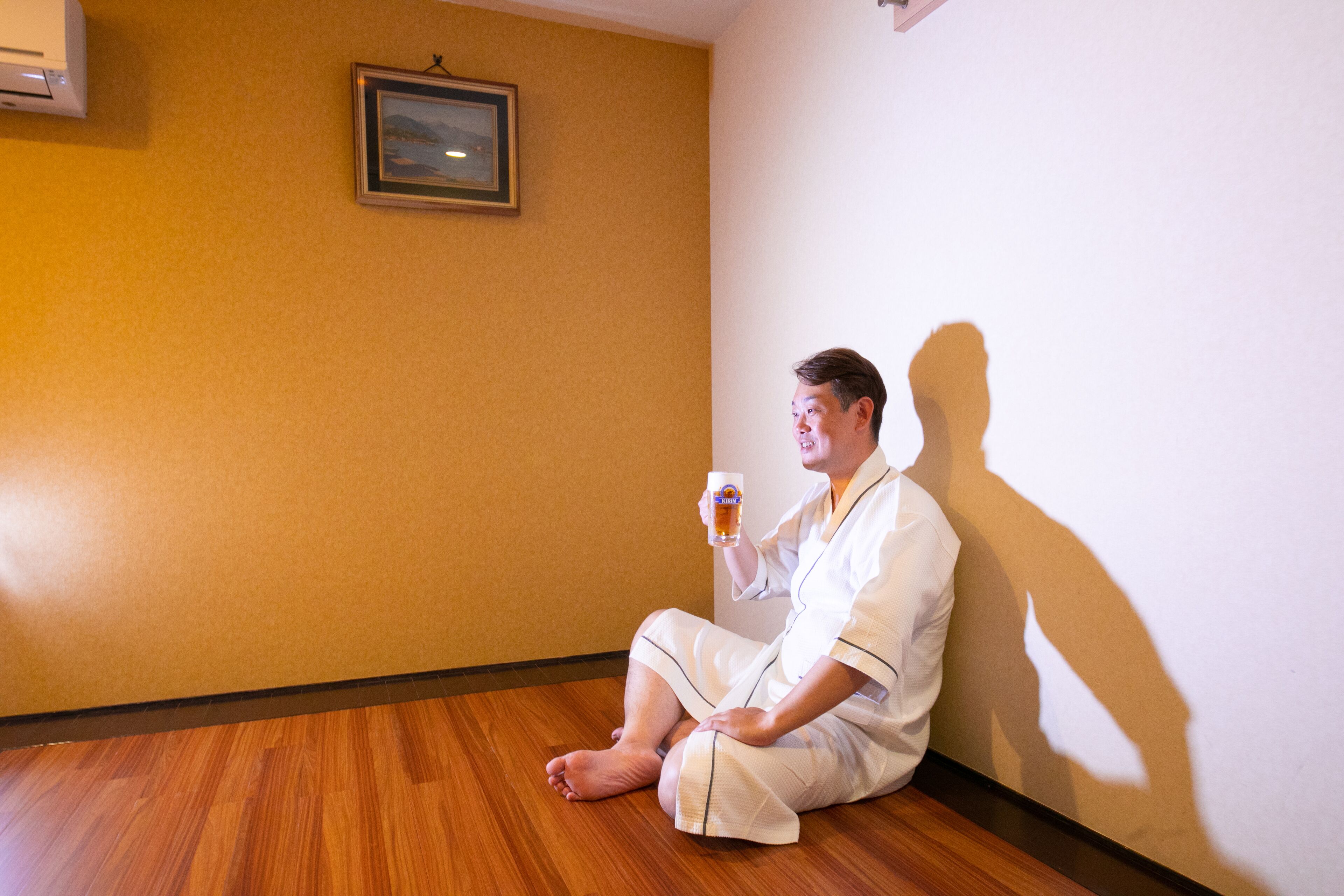 A person sitting on the floor in a room with wooden flooring and a plain wall.