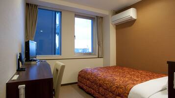 A hotel room with a desk, a bed with a patterned bedspread, a window with curtains, and an air conditioning unit.