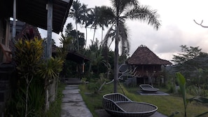 A pathway leading to a traditional wooden structure with a tiled roof, surrounded by tropical vegetation and a clear sky. - Kubu Taru (Sidemen)