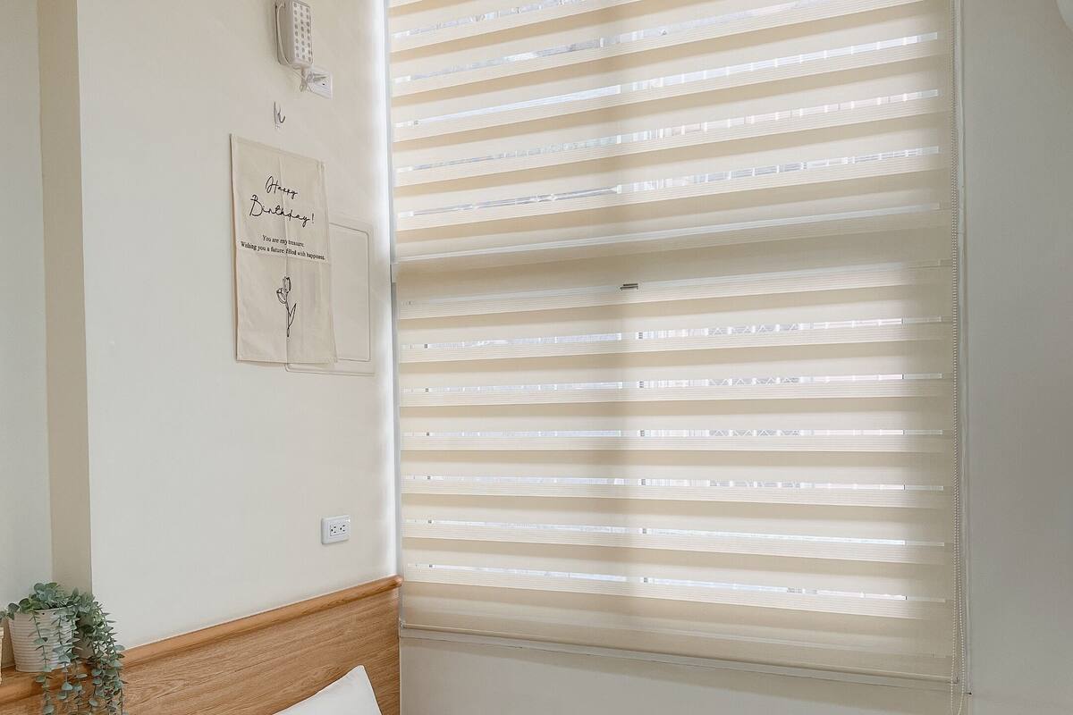 A single bed with white bedding, a wooden headboard, a red pillow, a wall-mounted air conditioner, and a window with horizontal blinds.