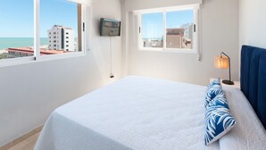 Et hotelværelse med en seng, et fjernsyn ophængt på væggen og udsigt til bygninger og havet gennem vinduerne.