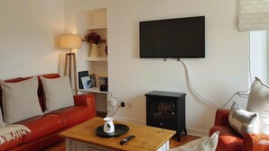 A living room with a red leather sofa, a wooden coffee table, a black wood-burning stove, a flat-screen TV mounted on the wall, and a window with blinds. - The Corner Cottage Sandwick (Sandwick)