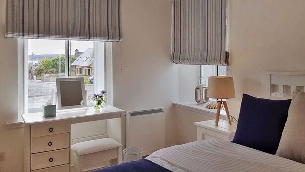 A bedroom with a bed, a bedside table with a lamp, a dresser with drawers, a window with a view of the street, and a radiator. - The Corner Cottage Sandwick (Sandwick)