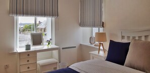 Un dormitorio con cama, una mesita de noche con lámpara, cómoda con cajones, ventana con vista a la calle y radiador.