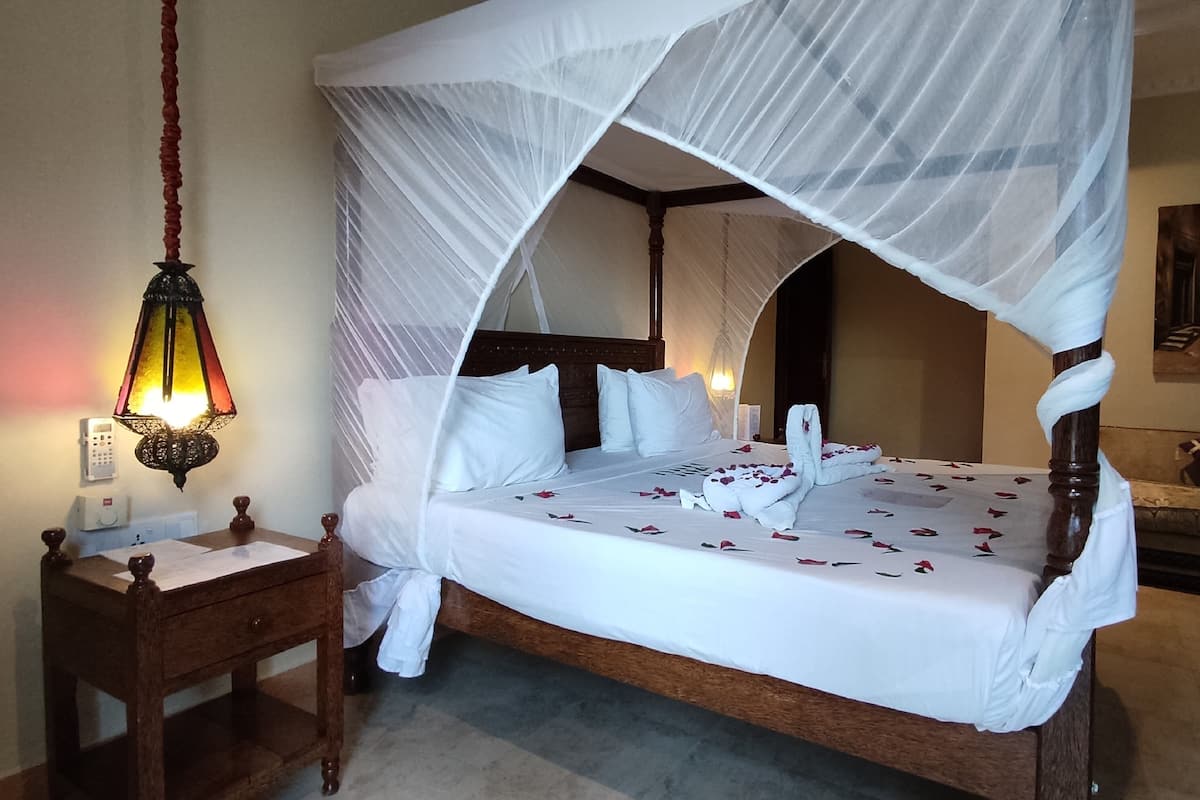 Una cama con dosel y cuatro columnas con sábanas blancas y pétalos de rosa, una mesita de noche con una lámpara, y un cuadro en la pared.