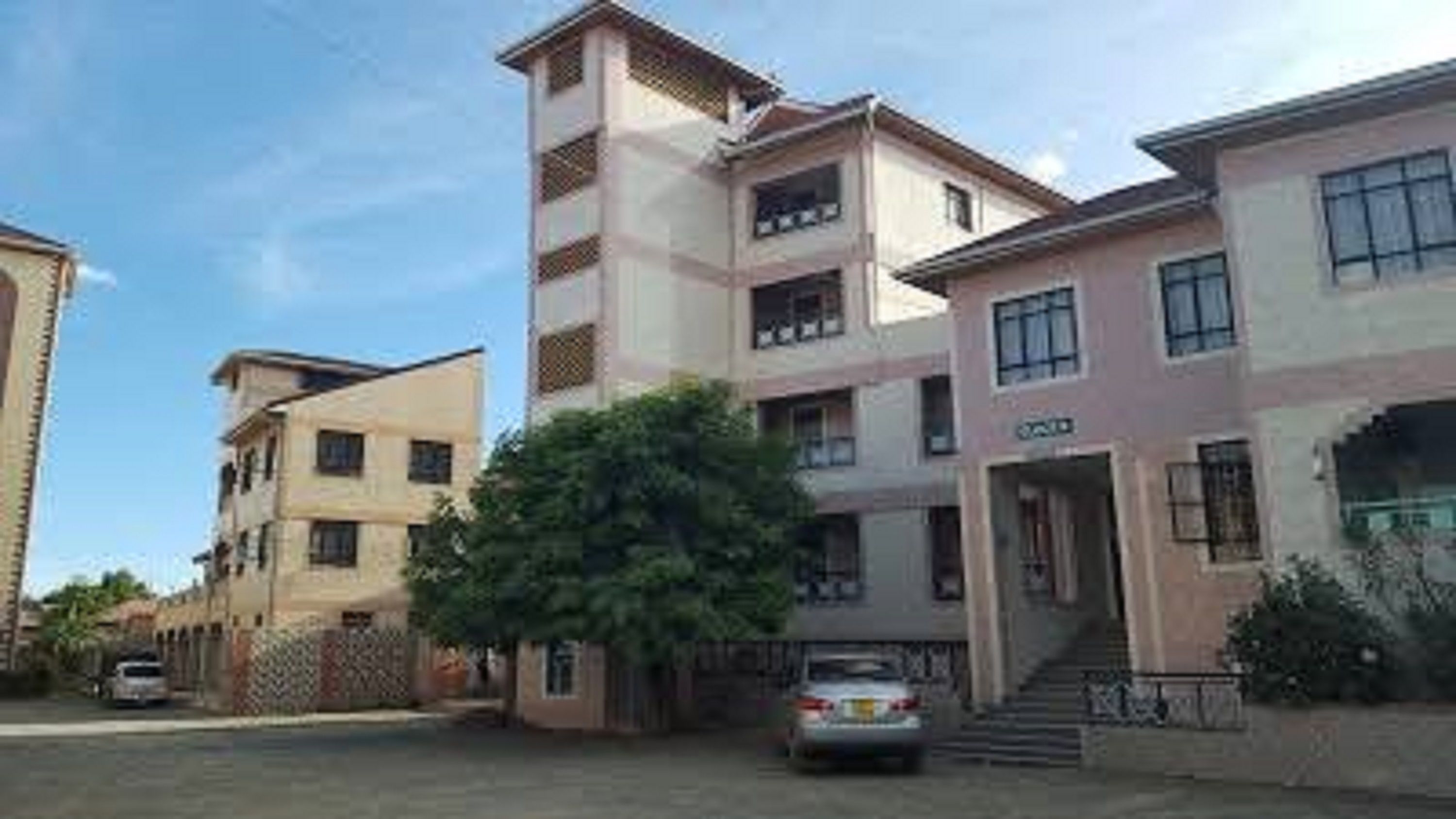 A multi-story residential building with a central staircase, a car parked in front, and a tree in the foreground.