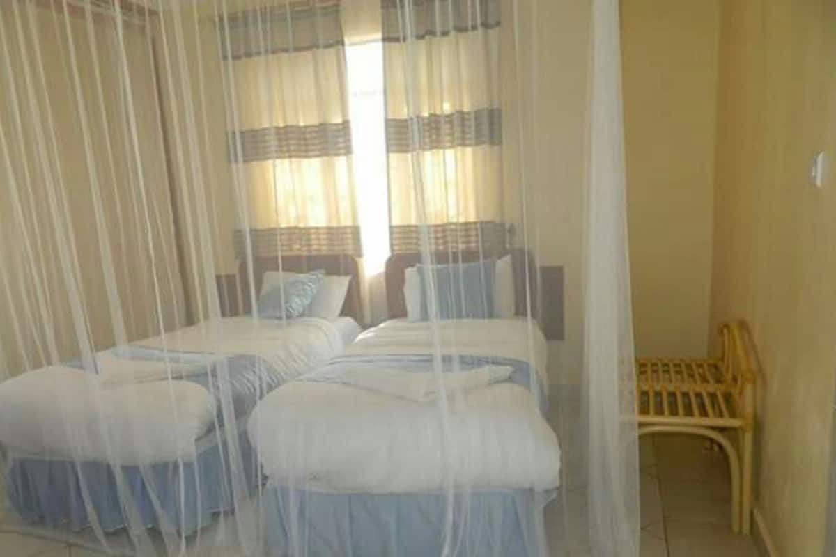 Quarto com duas camas cobertas por mosquiteiro, uma cadeira de madeira e uma janela com cortinas.