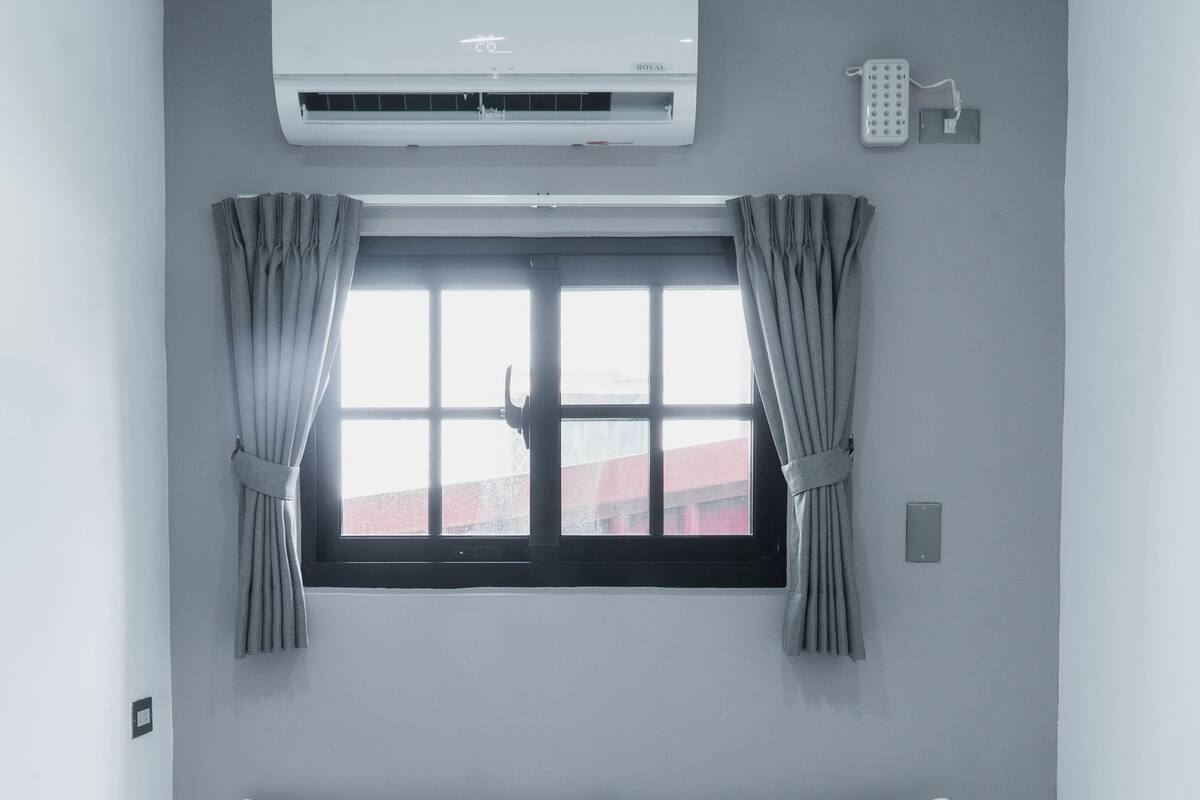 A hotel room with a bed, white bedding, a window with curtains, an air conditioning unit, and a wall-mounted phone.
