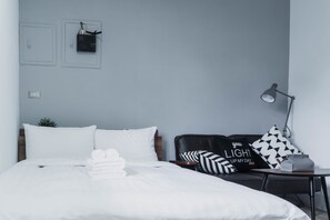 A neatly made bed with white linens, a black leather sofa with patterned cushions, a small table with a lamp, and a wall-mounted power outlet. - Beizhu Tainan Homestay (Tainan)