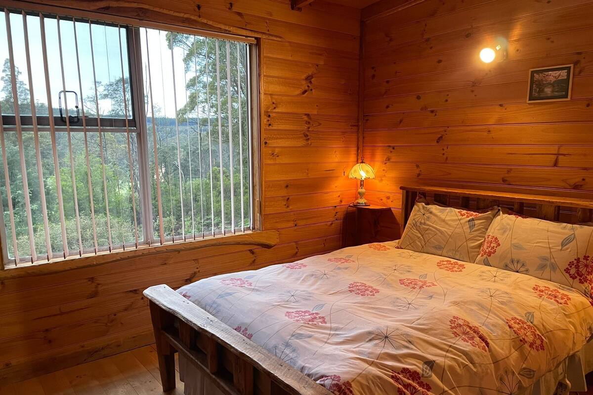 Una camera in una casetta di legno con un letto, un comodino, una finestra con persiane e un quadro appeso al muro.