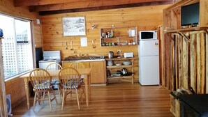 Interior de una cabaña de madera con una mesa de comedor, sillas, una zona de cocina con estufa, microondas y estanterías, y una chimenea.