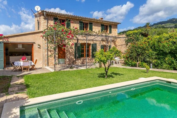 A two-story stone building with a swimming pool, a patio with outdoor furniture, and a lush garden.