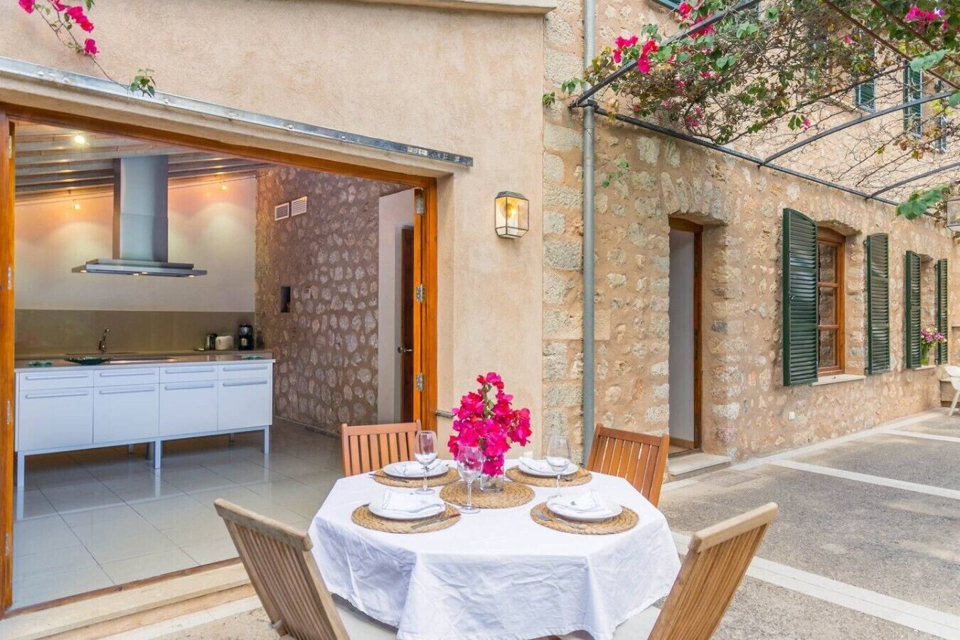 Un comedor con una mesa dispuesta para dos, una cocina visible a través de una puerta abierta, y un edificio de piedra con contraventanas verdes.