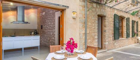 A dining area with a table set for two, a kitchen visible through an open door, and a stone building with green shutters.