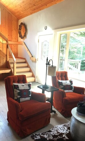 A wooden interior with a staircase, red armchairs, a lamp, and a view of greenery outside.
