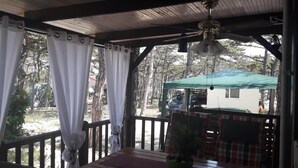 A covered outdoor seating area with a table and chairs, a ceiling fan, and a view of a house and trees. - Kamp Planik- Campsite (Razanac)