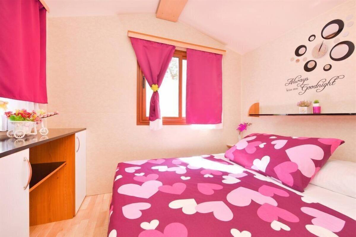 A bedroom with a bed covered in a pink and white heart-patterned blanket, a window with pink curtains, and a wall decoration with the phrase "Always kiss me Goodnight".