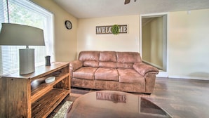 A living room with a leather couch, wooden coffee table, and a lamp. There is a 'WELCOME' sign on the wall and a clock.