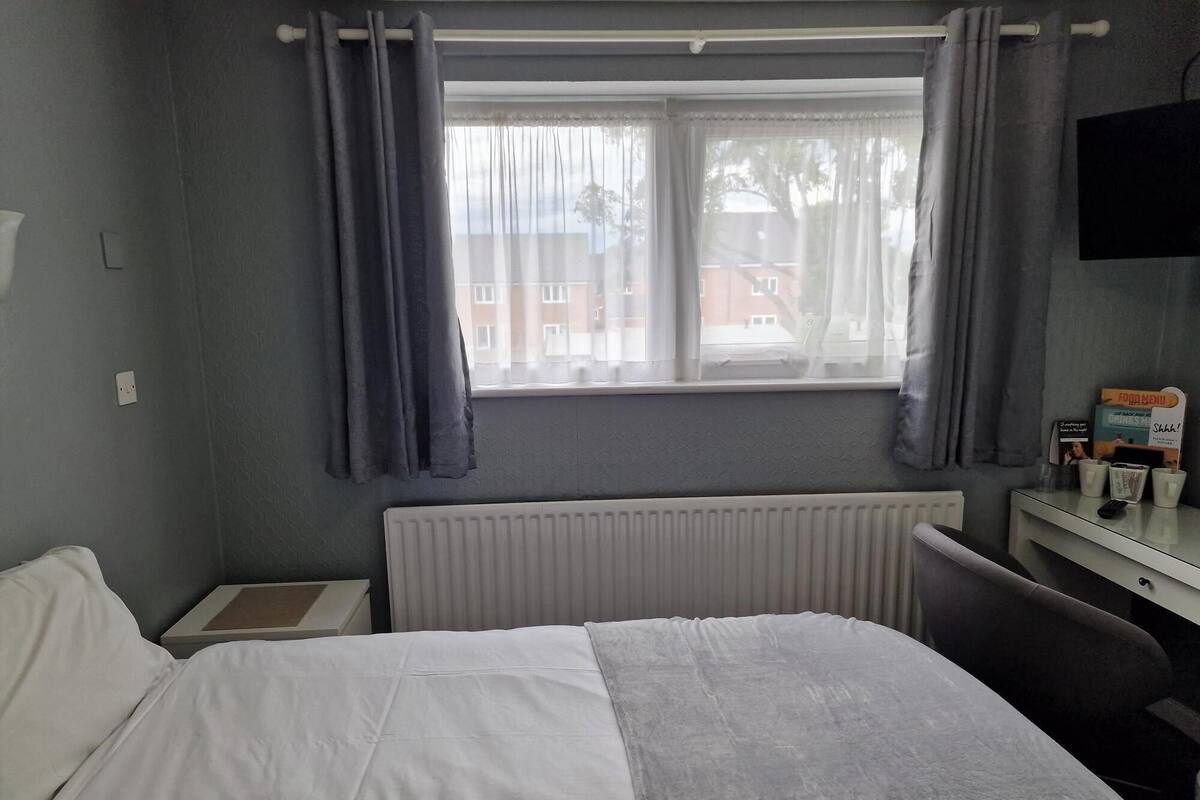 A hotel room with a bed, a desk, a chair, a window with curtains, a radiator, and a television on the wall.