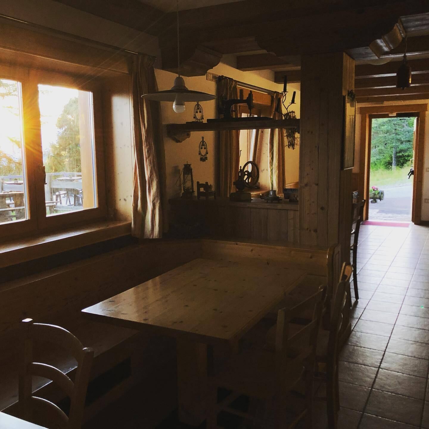 A rustic wooden interior with a large table and chairs, a window with curtains, and a view of the outdoors.