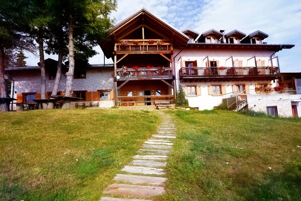 A two-story wooden building with balconies, a stone facade, and a stone pathway leading to the entrance.