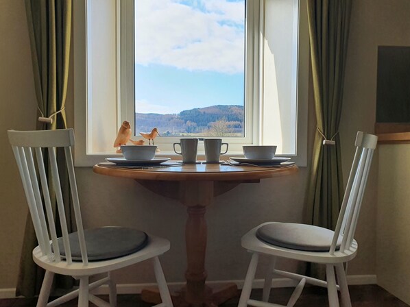 A dining table with two white chairs, a window with a view of a forest, and a small figurine of birds on the windowsill.