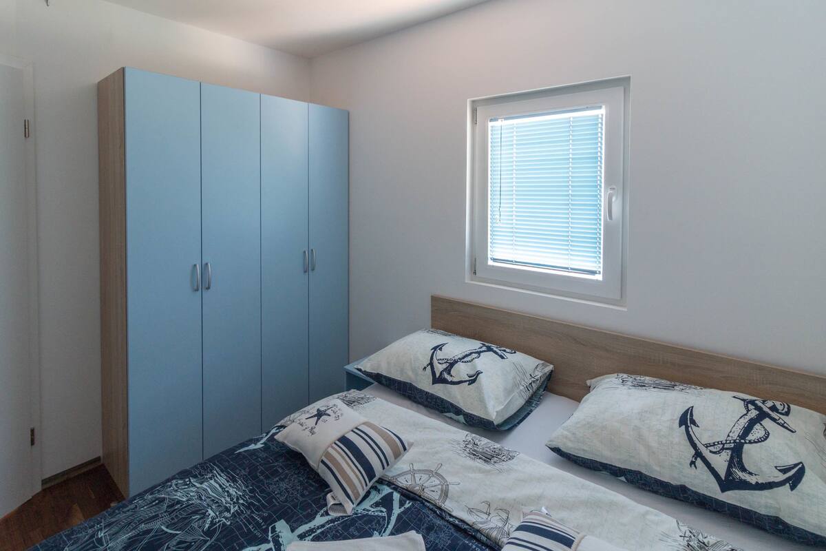 Une chambre à coucher avec un lit, deux oreillers ornés de motifs marins, une armoire et une fenêtre avec des stores.