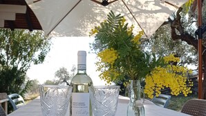 Una mesa dispuesta con una botella de Pinot Grigio, dos copas, un bloque de queso, una tabla de madera y un tarro con flores amarillas.