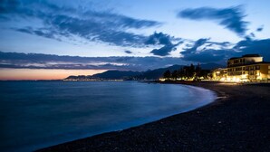 Escena costera con playa, un edificio con ventanas iluminadas y montañas al fondo.