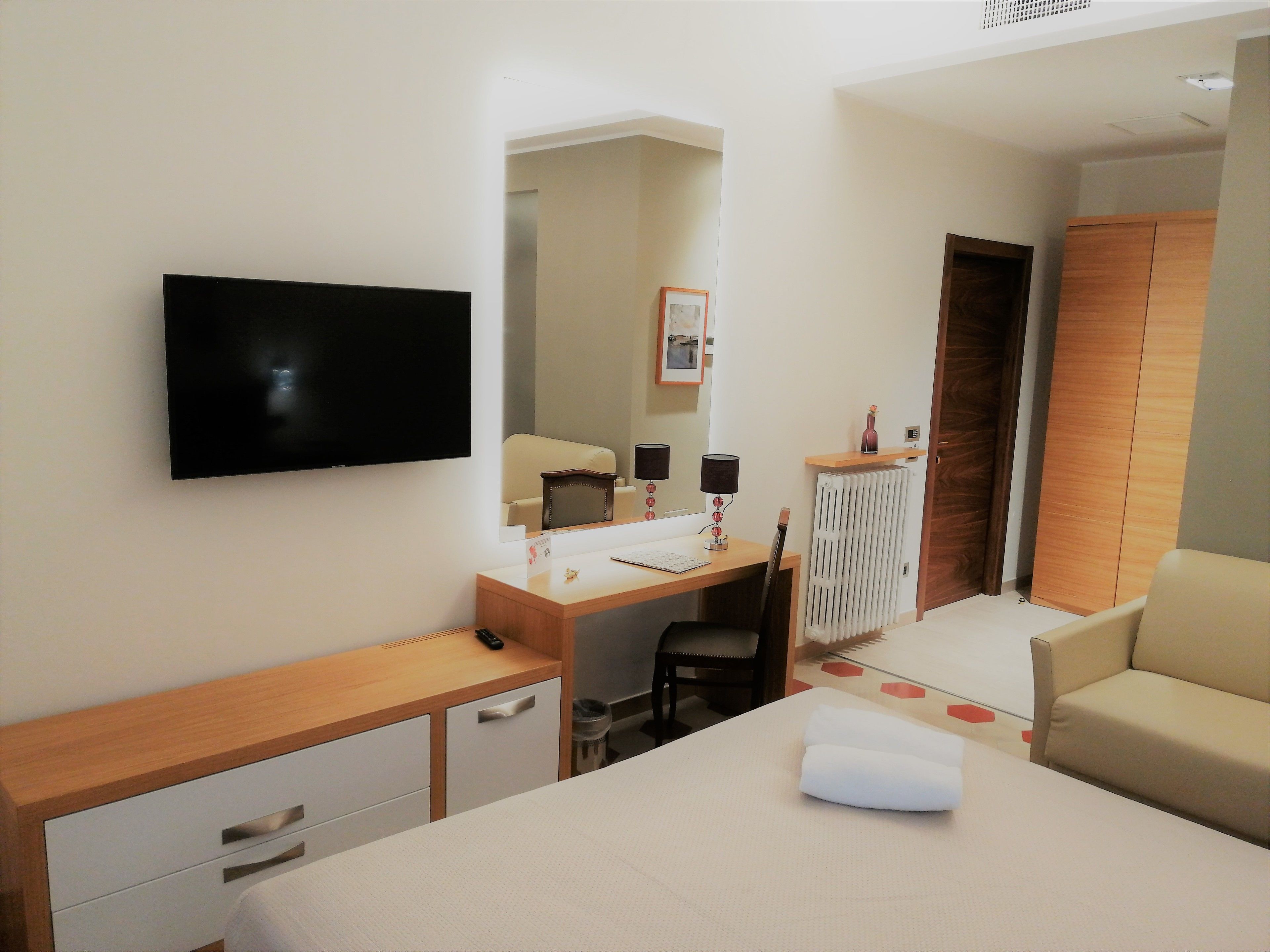A hotel room with a bed, a desk, a chair, a TV, a radiator, and a wardrobe.