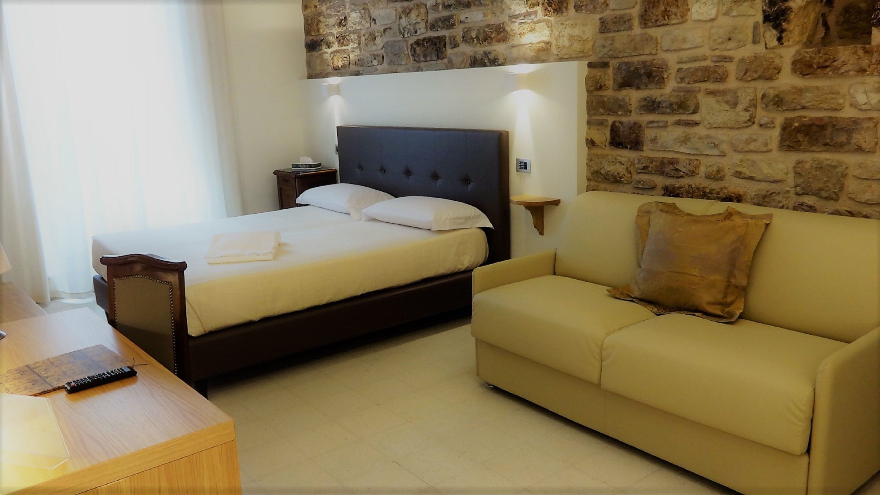 A hotel room with a bed, a sofa, a wooden desk, and a stone wall.