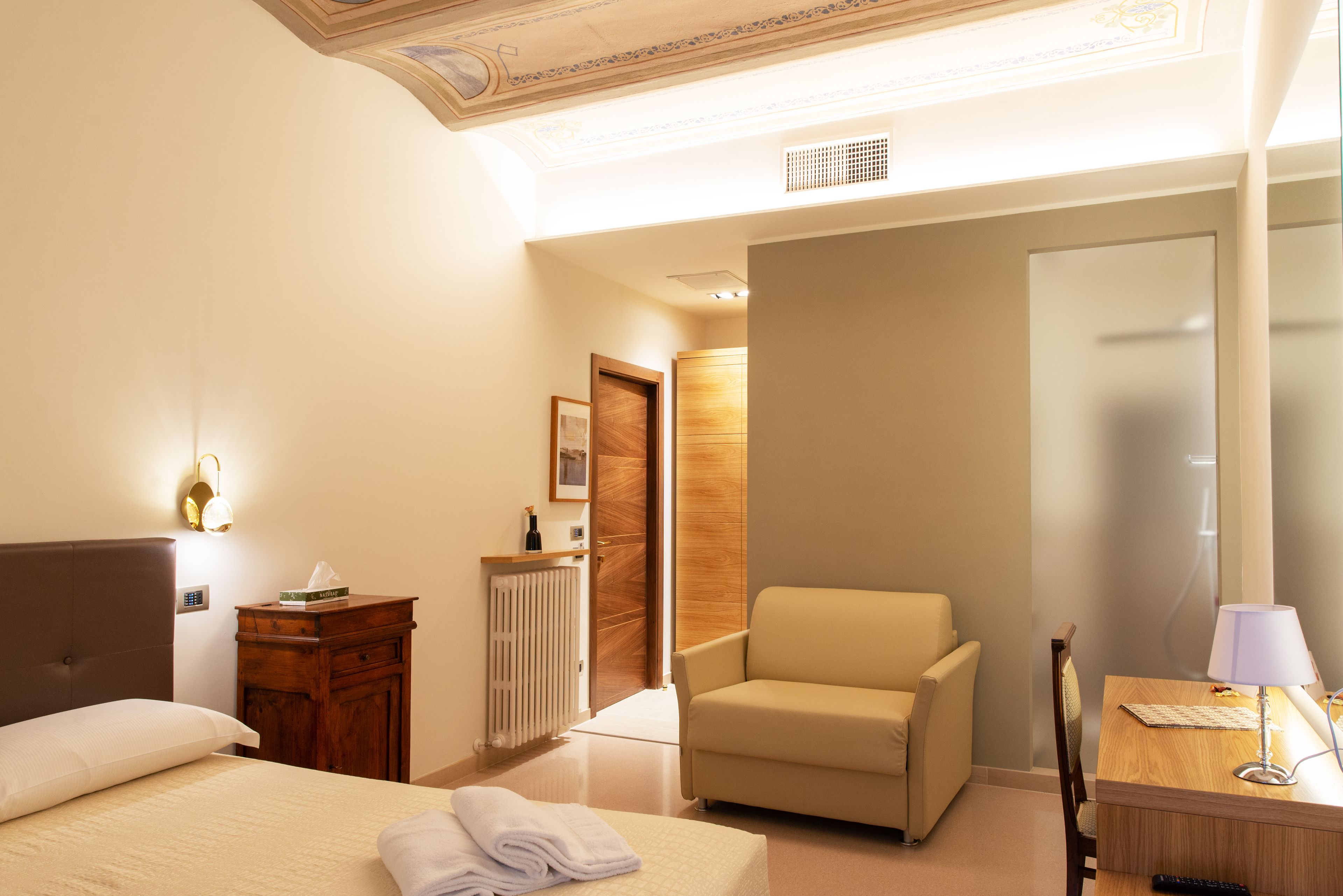 Habitación de hotel con una cama, una silla, un escritorio con lámpara, un radiador y una puerta que da a otra habitación.
