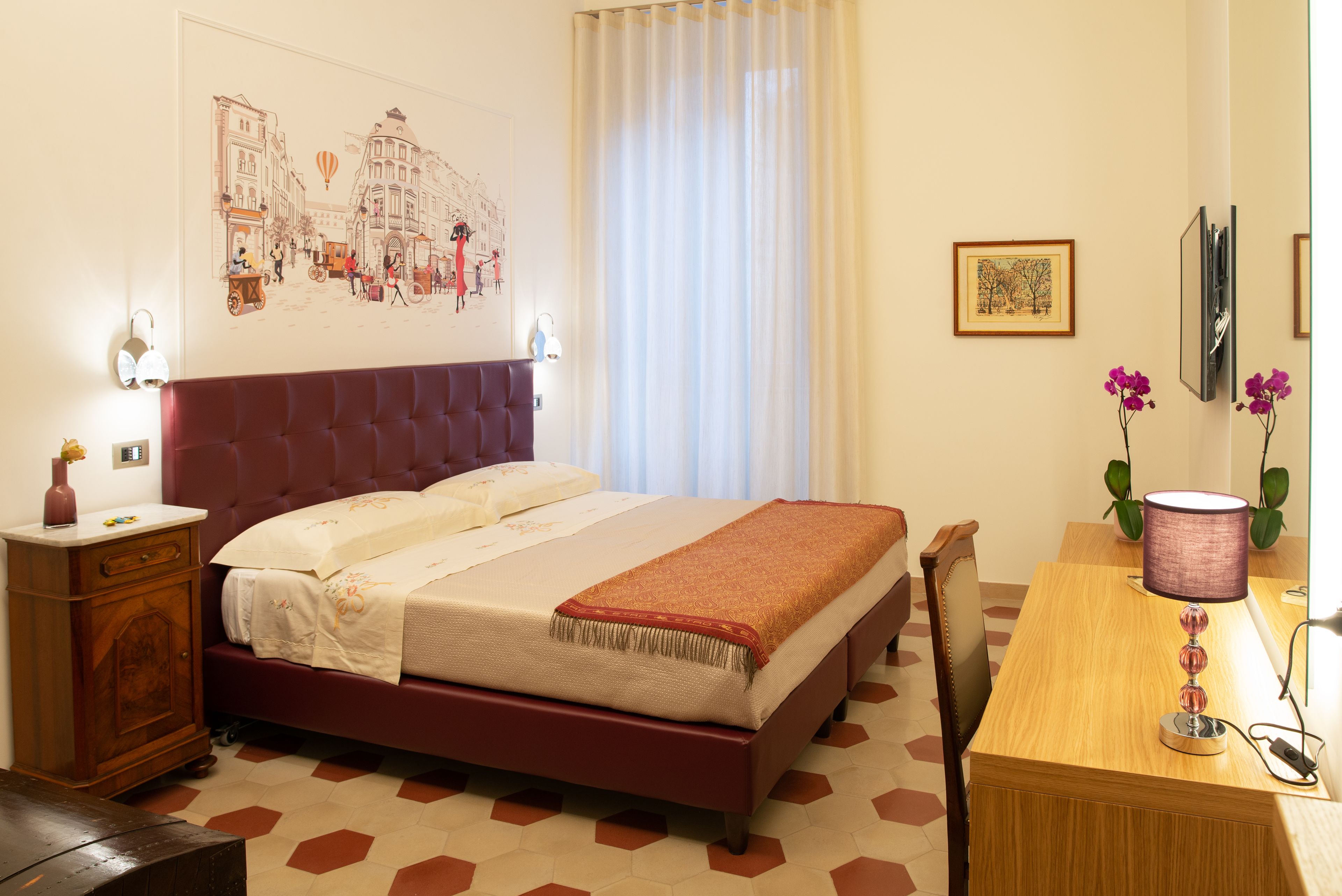 Une chambre d’hôtel comprenant un lit, un bureau en bois, une chaise, une table de chevet, une lampe et un mur orné d’une œuvre d’art représentant un paysage urbain.