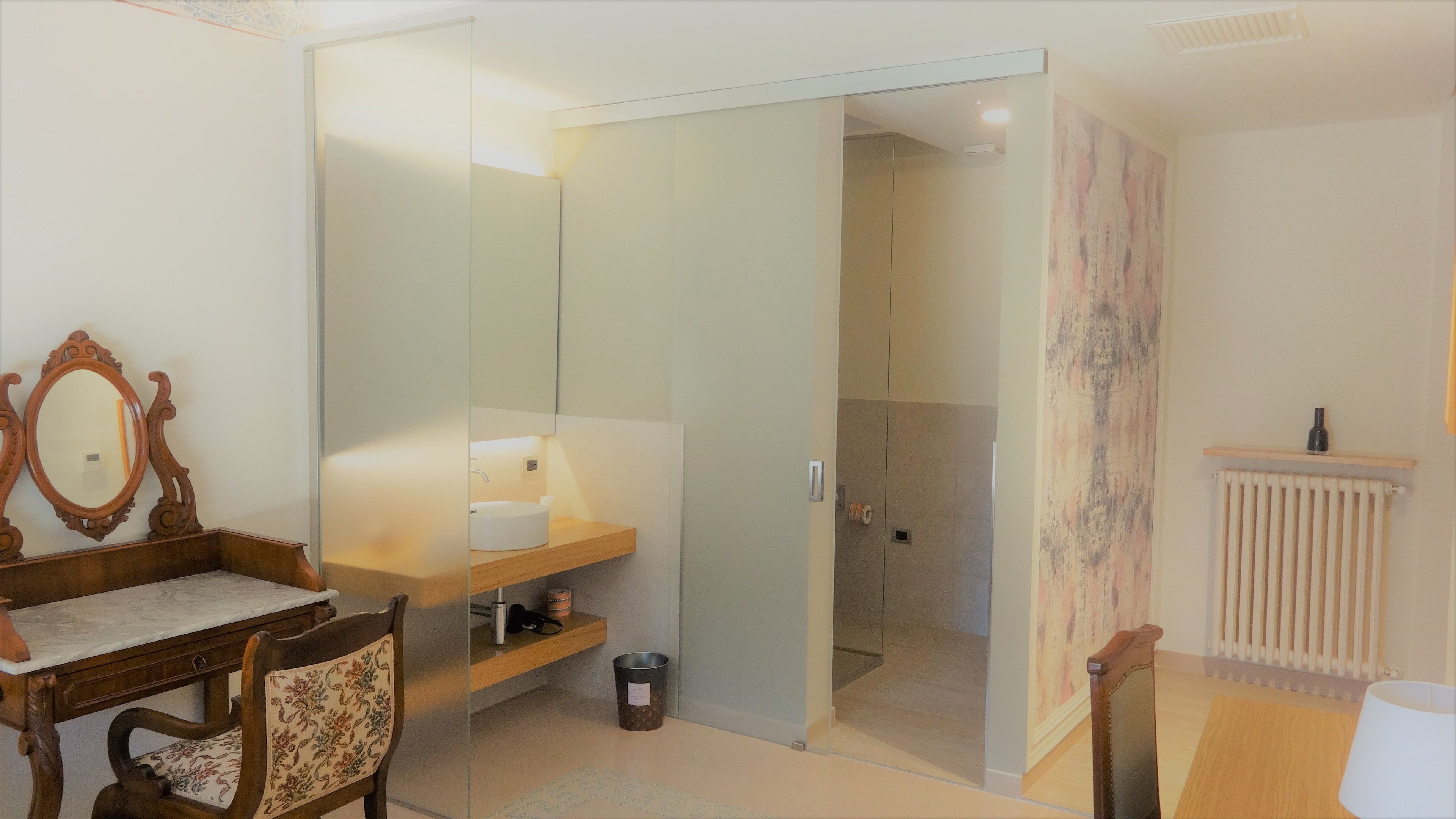 A room with a wooden desk, a chair, a mirror, a white sink, a radiator, and a door leading to another room.