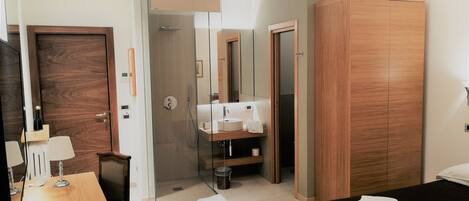 A modern hotel room with a wooden wardrobe, a glass-enclosed shower, a wooden desk with a lamp, and a bed with folded towels.