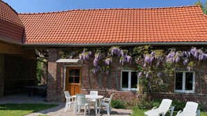 A brick building with a red-tiled roof, a wooden door, and a small patio with a table and chairs.