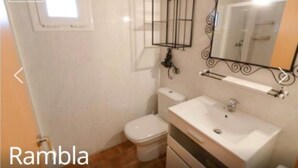 Ein Badezimmer mit Toilette, Waschbecken und Spiegel. Über der Toilette befindet sich ein Fenster und an der Wand ein Metallregal.