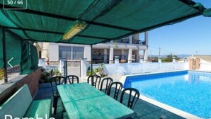 Ein Poolbereich mit Tisch und Stühlen, eine überdachte Sitzgelegenheit und ein Gebäude im Hintergrund.