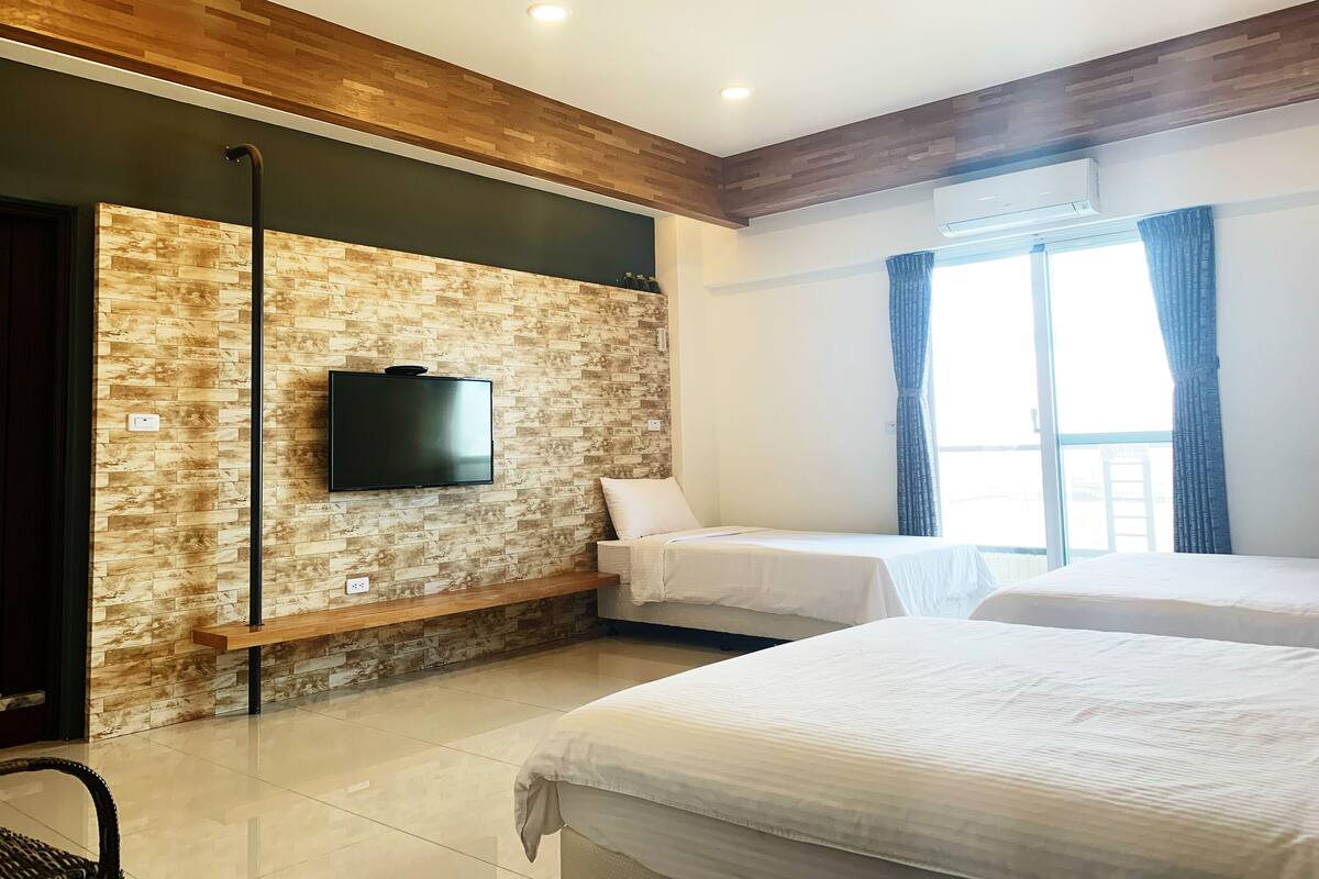 A hotel room with a brick-patterned wall, a flat-screen TV, a bed, a bench, and a window with curtains.