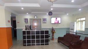 A reception area with a reception desk, a television mounted on the wall, a clock, and a seating area with brown chairs. - Galpin Suites Ikeja (Lagos)