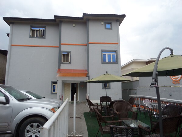 A building with a grey facade, orange accents, and multiple windows. There are outdoor wicker chairs and tables under umbrellas, and a car parked in front. - Galpin Suites Ikeja (Lagos)