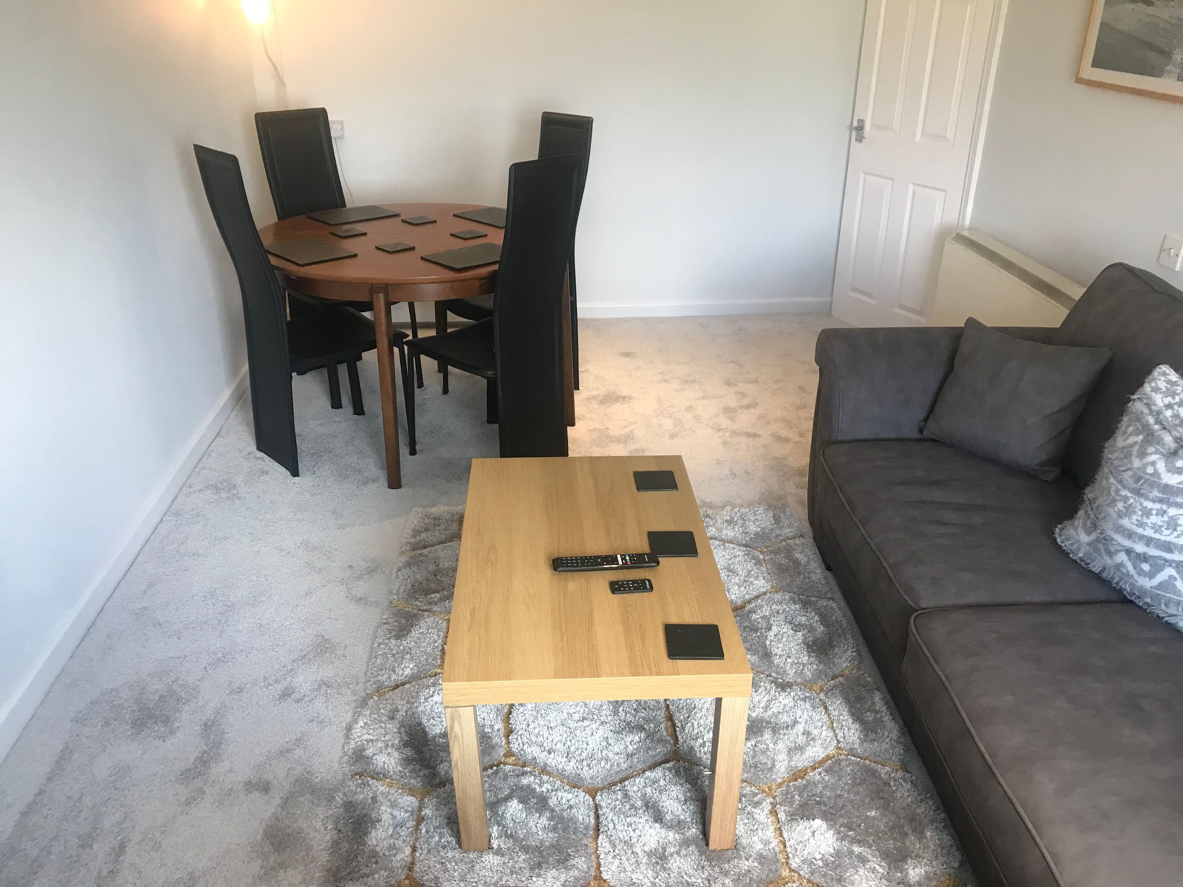 A living room with a wooden coffee table, a grey sofa, a dining table with chairs, and a patterned rug.