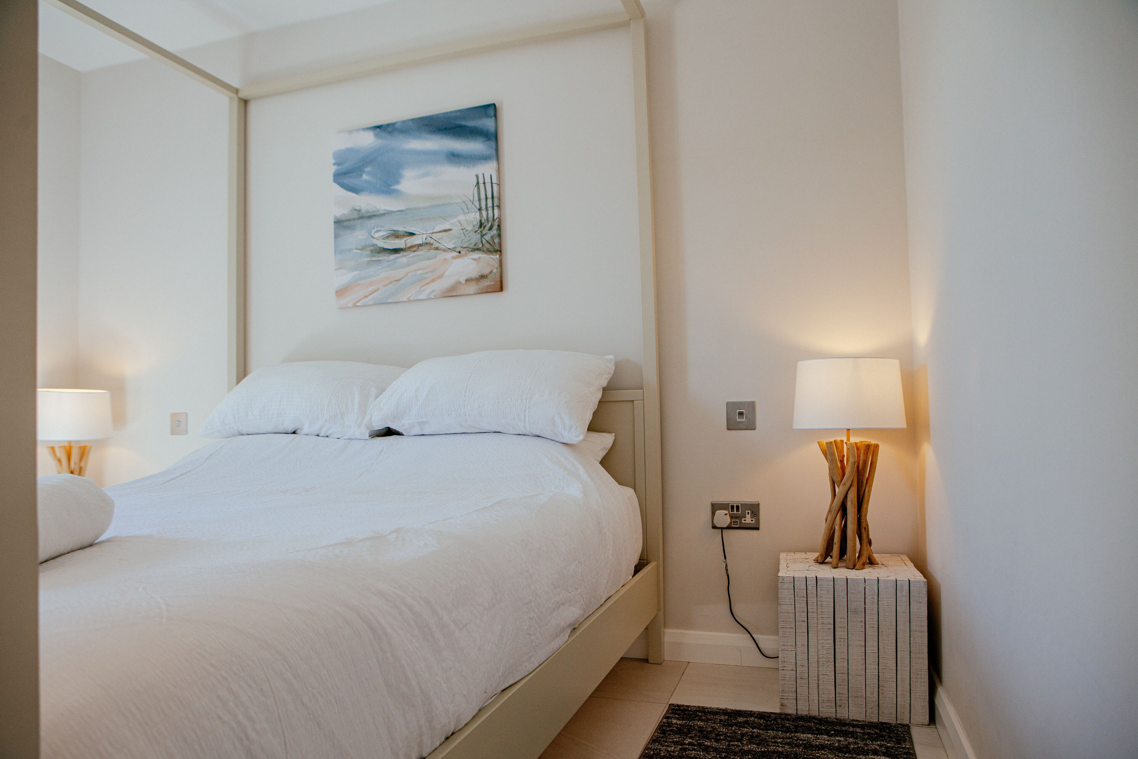 A neatly made bed with white linens in a room with a painting on the wall, a bedside lamp, and a power outlet.