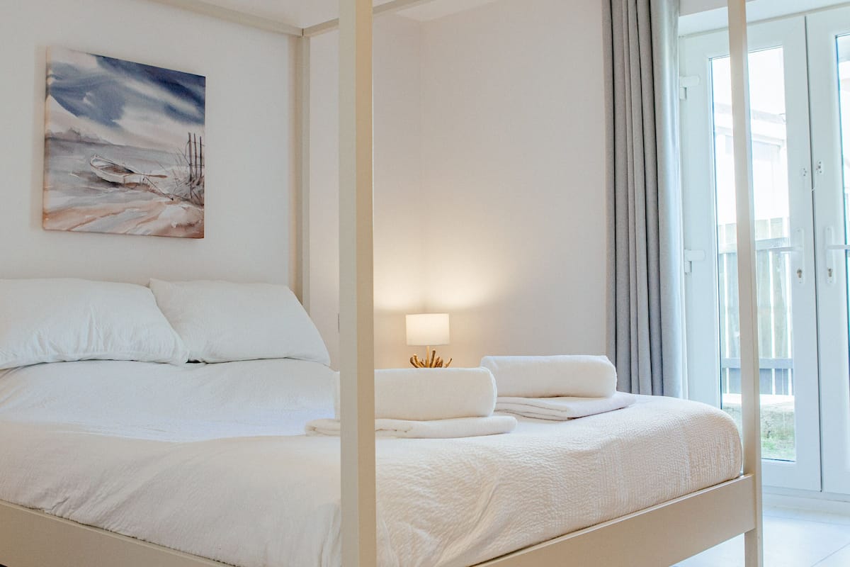 A four-poster bed with white bedding, a bedside lamp, and a painting on the wall.