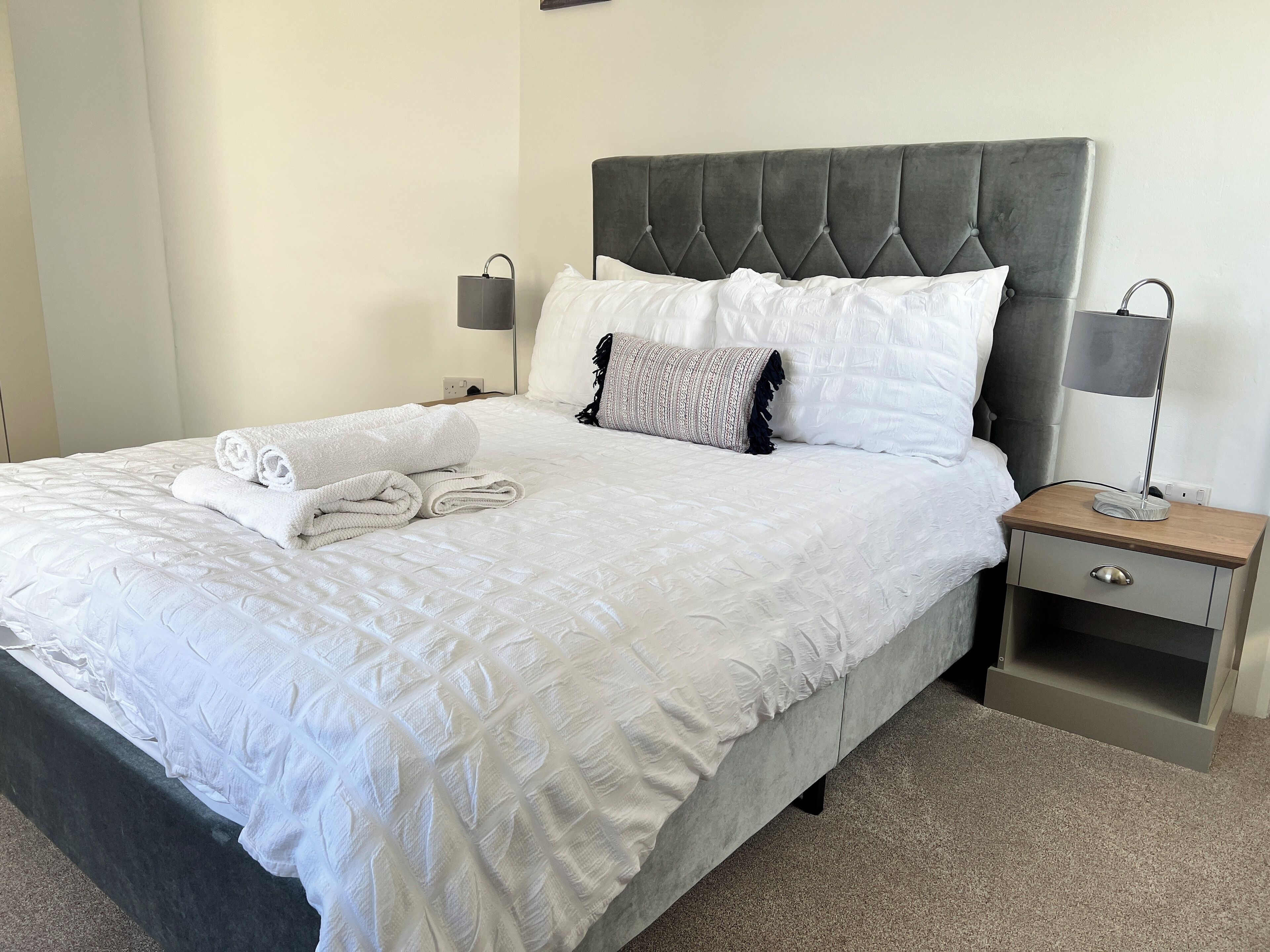 A neatly made bed with a quilted white duvet, two pillows, and a folded towel on the side. There is a bedside table with a lamp and a drawer.