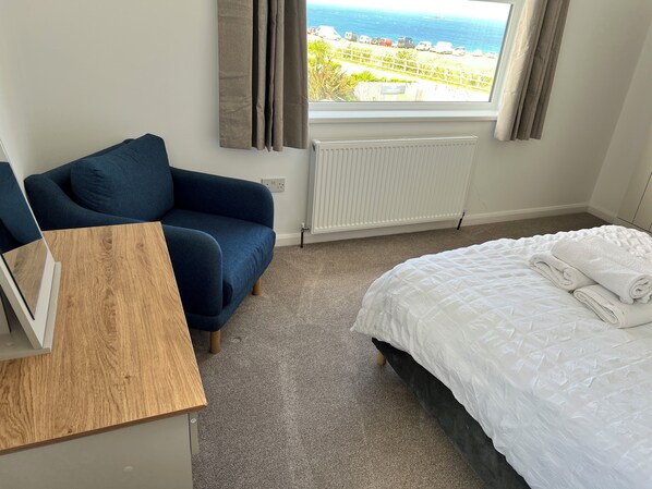 A hotel room with a bed, a chair, a wooden table, and a view of the sea through a window.