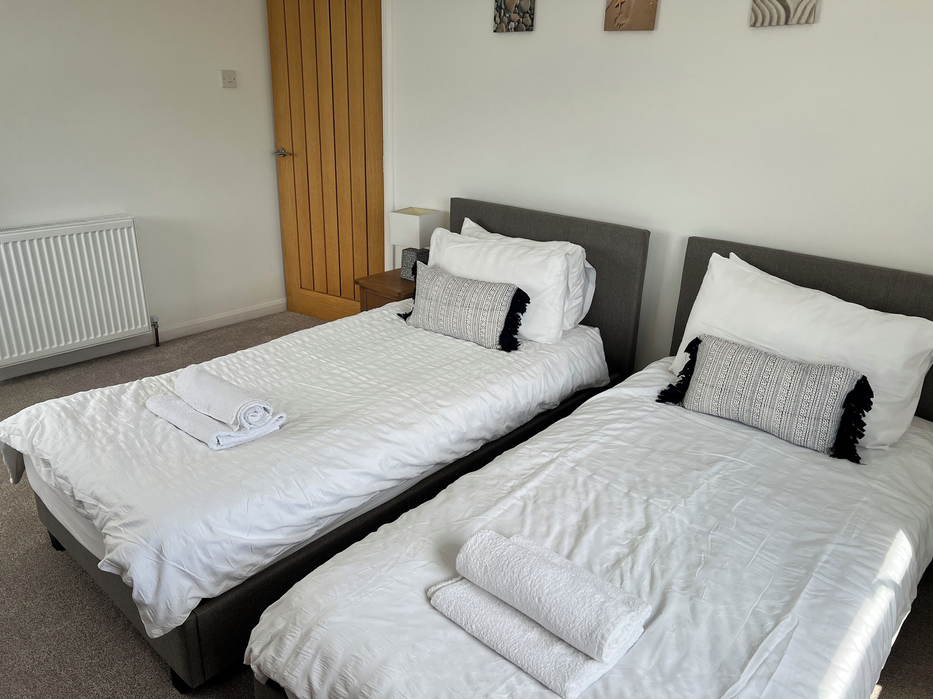 A room with two single beds, each with white bedding and a rolled towel. There is a wooden wardrobe and a radiator on the wall.